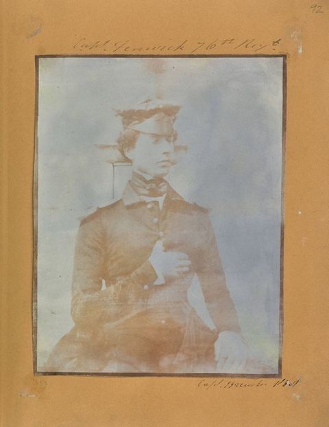 Black and white photograph. Shown from the lap up, a clean-shaven man wearing a uniform sits straight-backed, one thumb hooked between buttons of his coat so that hand lies flat against his chest.
