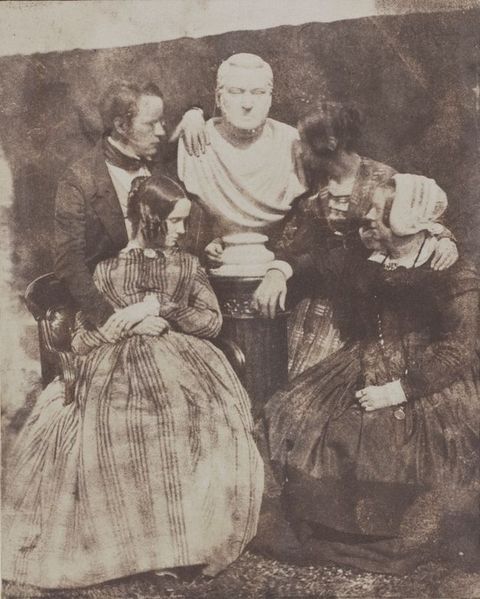 Black and white photograph. Three women and a man, all pale-skinned, gather around a marble bust of another man. One woman wearing a white bonnet rests a forearm on the pedestal base of the bust, and a younger woman wraps one hand around the backside of the base. The living man drapes one arm over the bust’s shoulder and rests the other on a young woman’s lap, who sits in front of him.