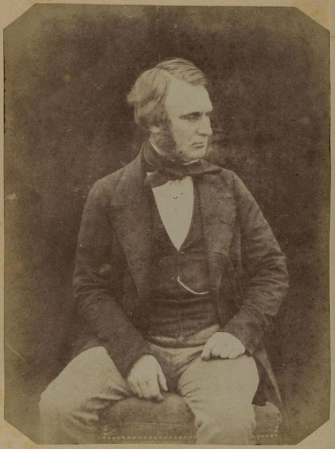 Black and white photograph. A pale-skinned man with mutton chops sits and looks off to our right. One hand rests at the top of his thigh, the other on the upholstered seat between his legs.