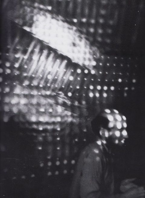 Black and white photograph. A person may be lit by holes in perforated material that also shine on the surface behind him, or may stand in front of perforated material in this vertical photograph.