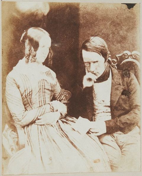 Black and white photograph. Shown from the laps up, a pale-skinned man and woman, both with dark hair, sit together. The man rests his chin in one hand, pince-nez balanced on the bridge of his nose as he reads a letter. The woman crosses her wrists, leans back in her seat, and gazes off to our right.