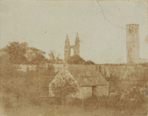 Black and white photograph. In tones of bronze and fawn brown, a small structure sits in front of a wall the same height as its pitched roof. A tower and ruins of a cathedral are outlined against the blank sky in the town beyond the wall.