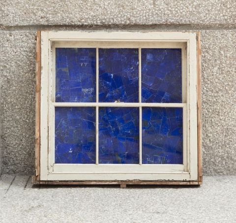 Sculpture. The openings of a six-paned window painted off-white are filled with pieces of lapis lazuli, so they practically glow blue. The window leans up against a stone or concrete wall.