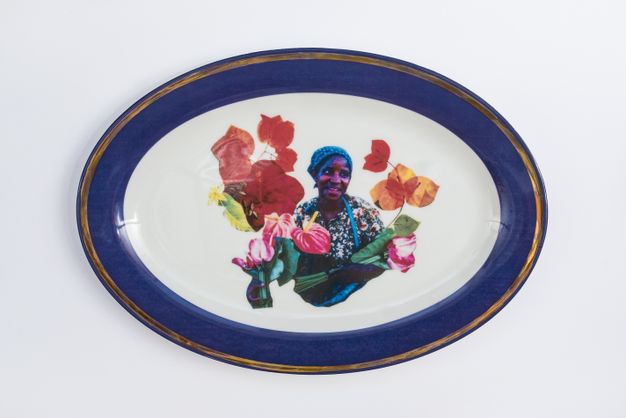 Porcelain. A dark-skinned woman looks off to our right and smiles at the centre of this white oval plate. She is surrounded by flowers and leaves. The cobalt-blue rim is edged in gold.