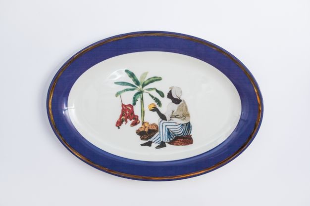 Porcelain. A dark-skinned woman sits near a palm tree holding yellow fruit in one hand at the centre of this white oval plate. She wears a white head covering and shirt, and blue-and-white striped, wide-legged pants. The cobalt-blue rim is edged in gold.